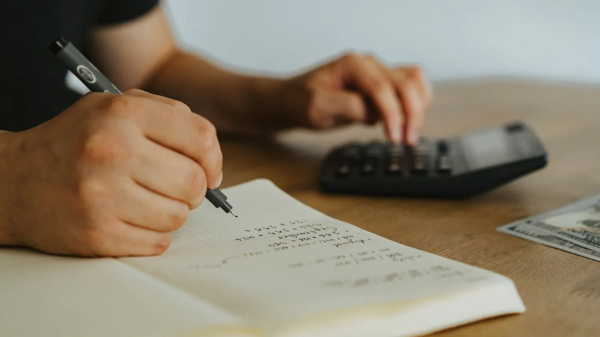 Mano anotando cuentas en un cuaderno junto a una calculadora