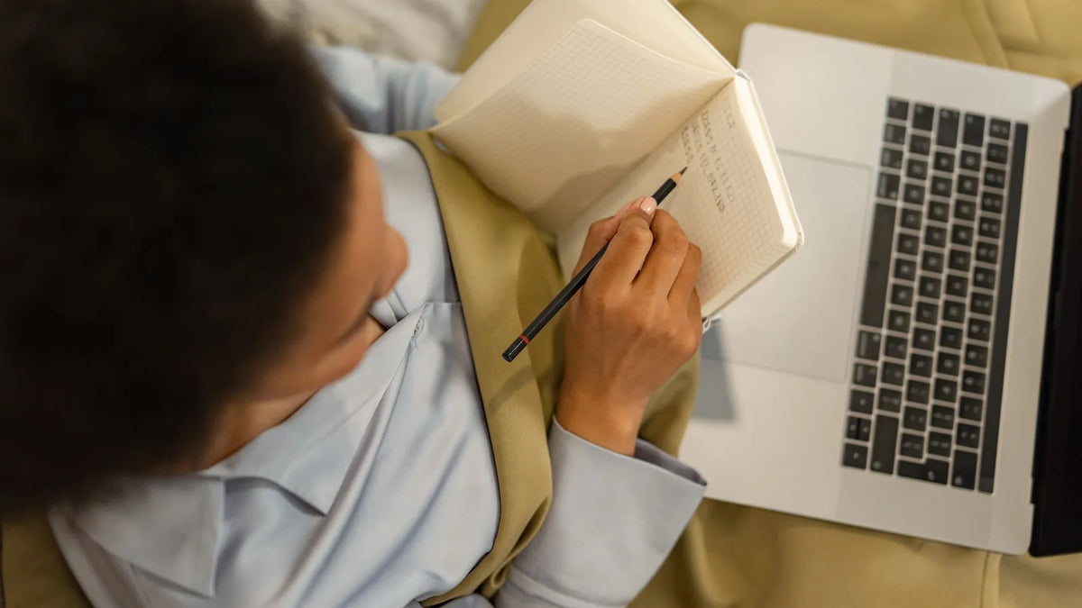 Persona tomando notas en un cuaderno con una laptop abierta sobre las piernas