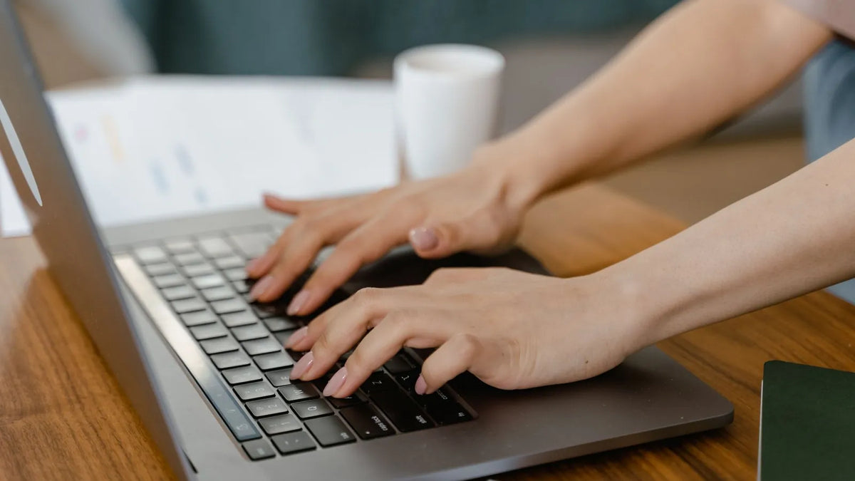 Persona escribiendo en una laptop con un cuaderno y una taza de café sobre un escritorio sencillo