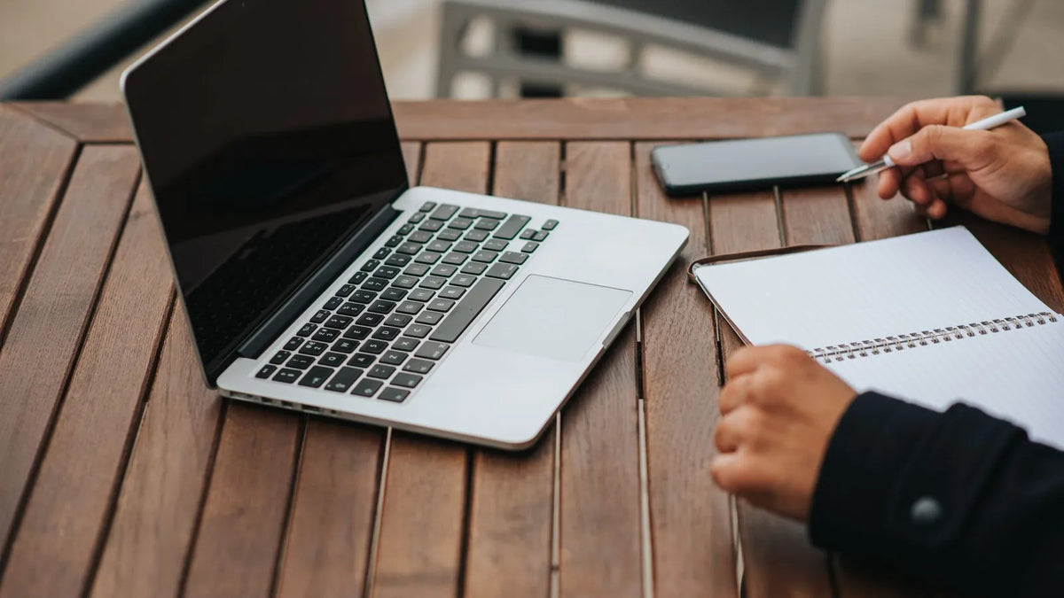 Laptop abierta y una persona anotando en un cuaderno sobre una mesa de madera