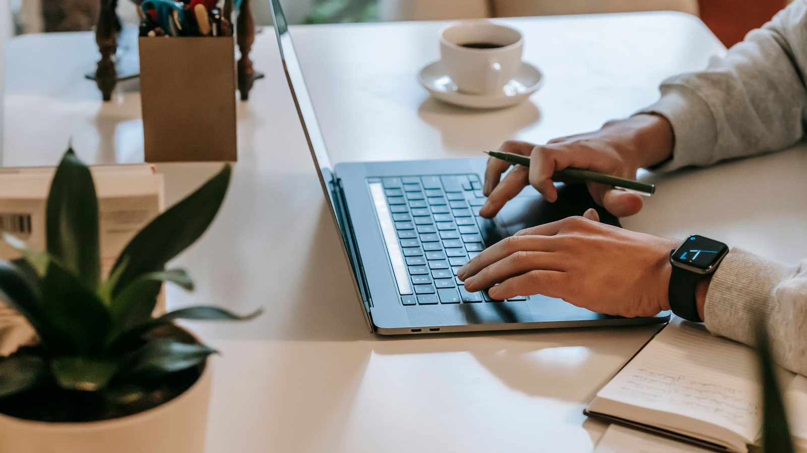 Manos escribiendo en una laptop junto a una taza de café y una planta