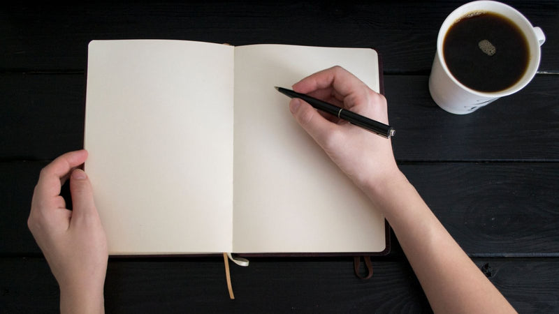 Mano escribiendo en una libreta en blanco junto a una taza de café