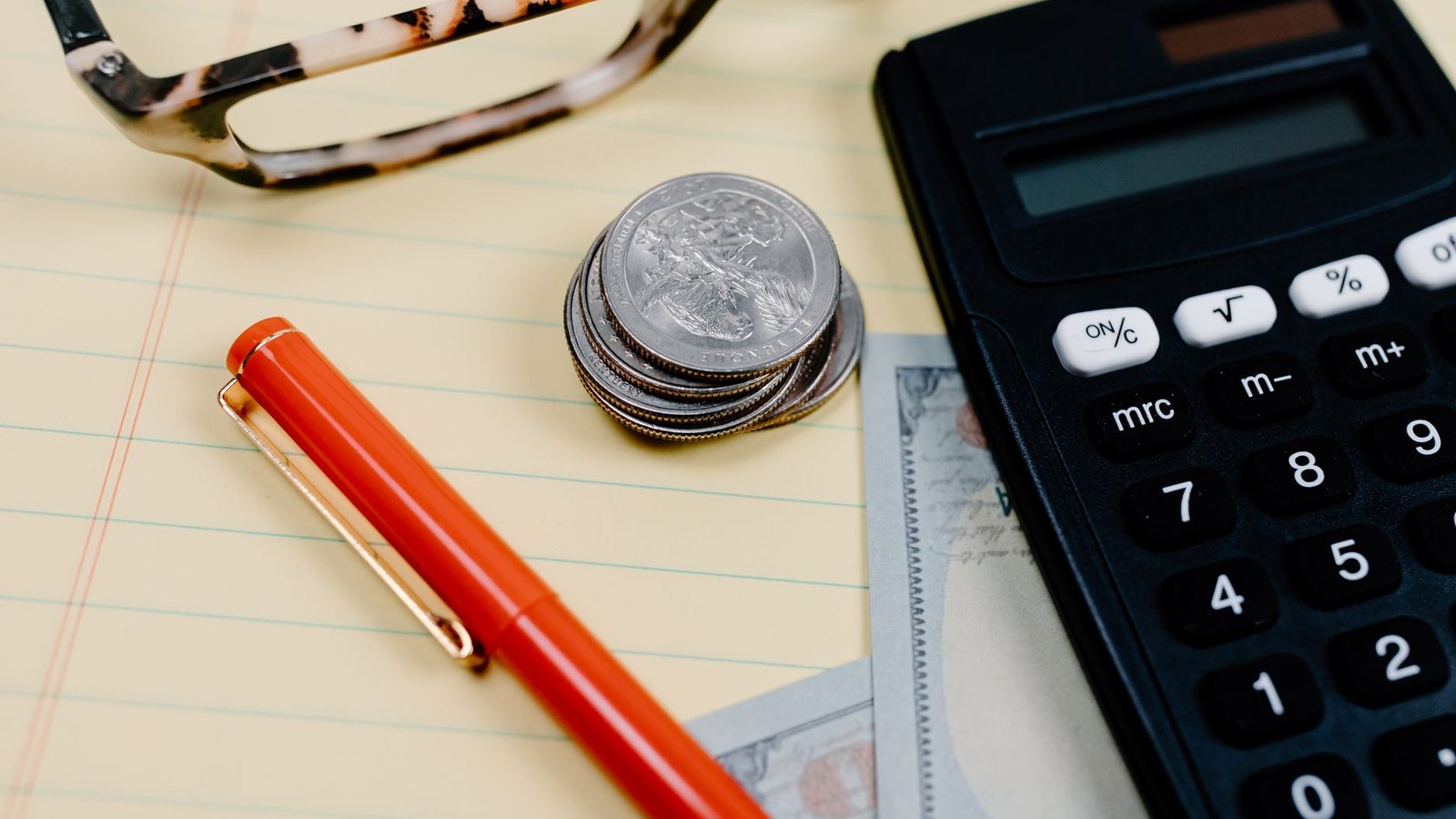 Calculadora, monedas, un bolígrafo rojo y unos lentes sobre un escritorio